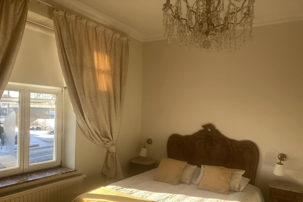 Een slaapkamer met een groot bed, een kroonluchter, een raam met gordijnen en een radiator.