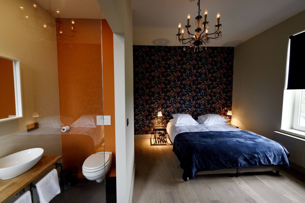 Een moderne slaapkamer met een groot bed, nachtkastje, een kroonluchter en een badkamer met toilet en wastafel.