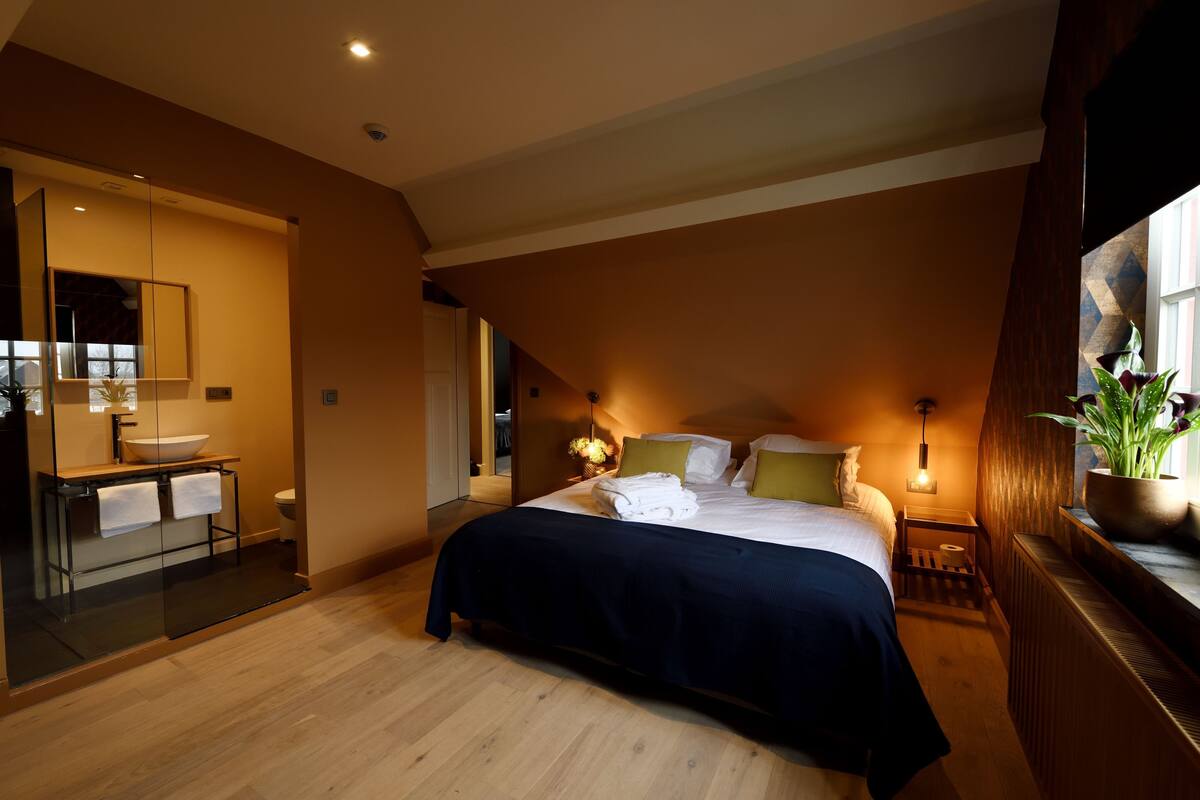 Een slaapkamer met een groot bed, nachtkastjes, een spiegel, een raam met uitzicht en een badkamer met een wastafel en spiegel.