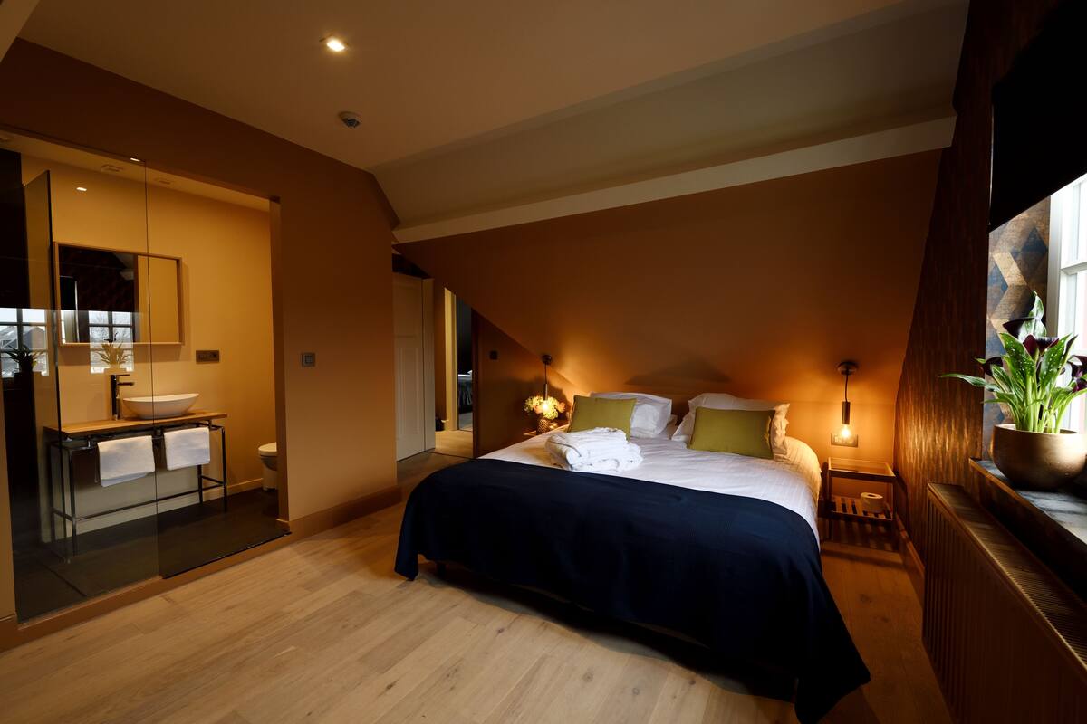 Een slaapkamer met een groot bed, nachtkastjes, een spiegel en een raam met een plant.