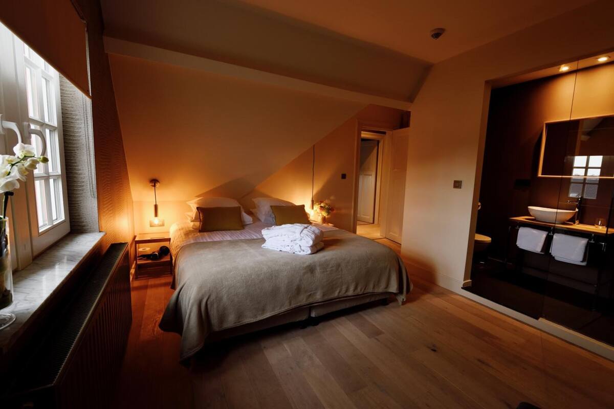 Een slaapkamer met een bed, nachtkastjes, een raam met gordijnen, en een badkamer met een spiegel en wastafel.
