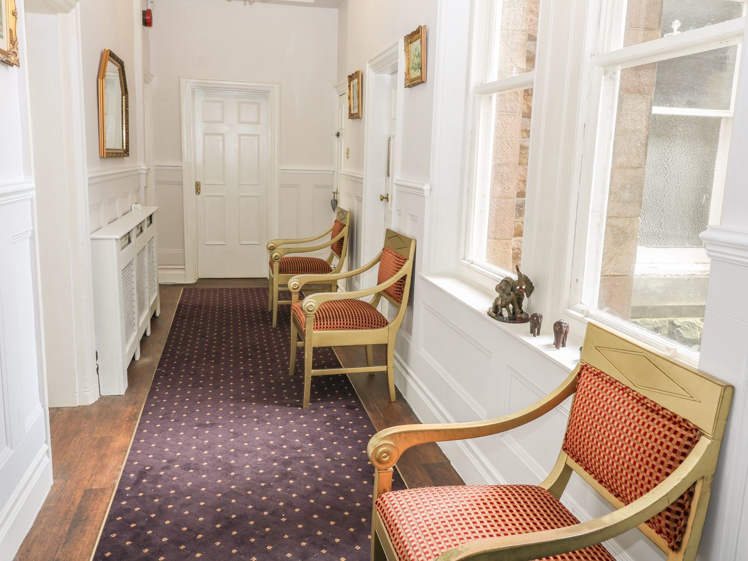 A hallway with a patterned carpet, wooden chairs with red cushions, a white door, and a window with a stone wall.