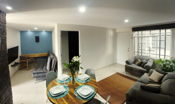 A modern living room with a dining area, a grey sofa, a TV mounted on a stone wall, and a large window with bars. - Hospedaje Tlaquepaque (Tlaquepaque)