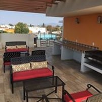 Un espacio de estar al aire libre con sillas metálicas y cojines rojos, una parrilla de barbacoa y vistas a la ciudad.