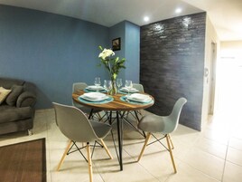 Un comedor con una mesa redonda dispuesta para seis personas, rodeada de sillas grises con patas de madera. Un jarrón con flores blancas está sobre la mesa. La habitación tiene paredes azules, una pared de acento de piedra y un sofá gris al fondo.