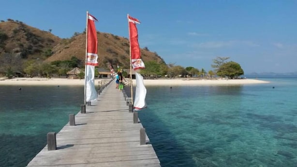 A wooden pier extending into clear turquoise water, flanked by red and white flags, leading to a sandy beach with sparse vegetation and a distant hill. - Situju 7 Cruises (Labuan Bajo)