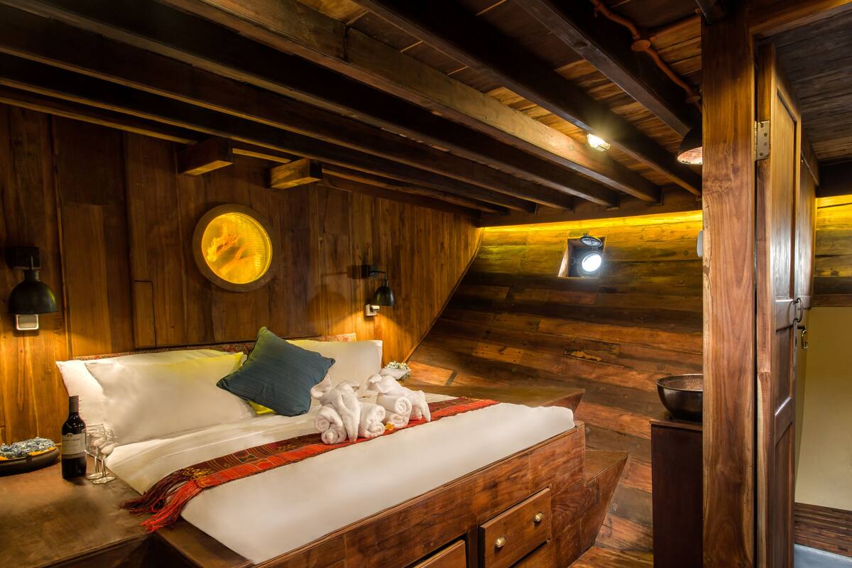 A cozy, rustic bedroom with a wooden ceiling, a bed with white linens, a round mirror, and two wall-mounted lamps.