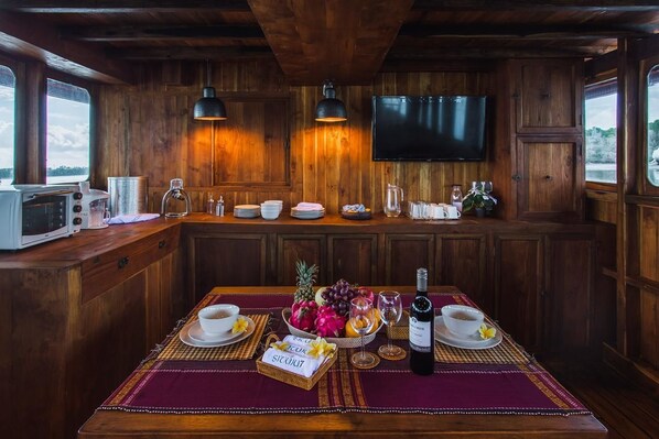 A wooden cabin interior with a kitchen area, a dining table set with a fruit bowl, wine glasses, and a bottle. A television is mounted on the wall above the kitchen counter. - Situju 7 Cruises (Labuan Bajo)