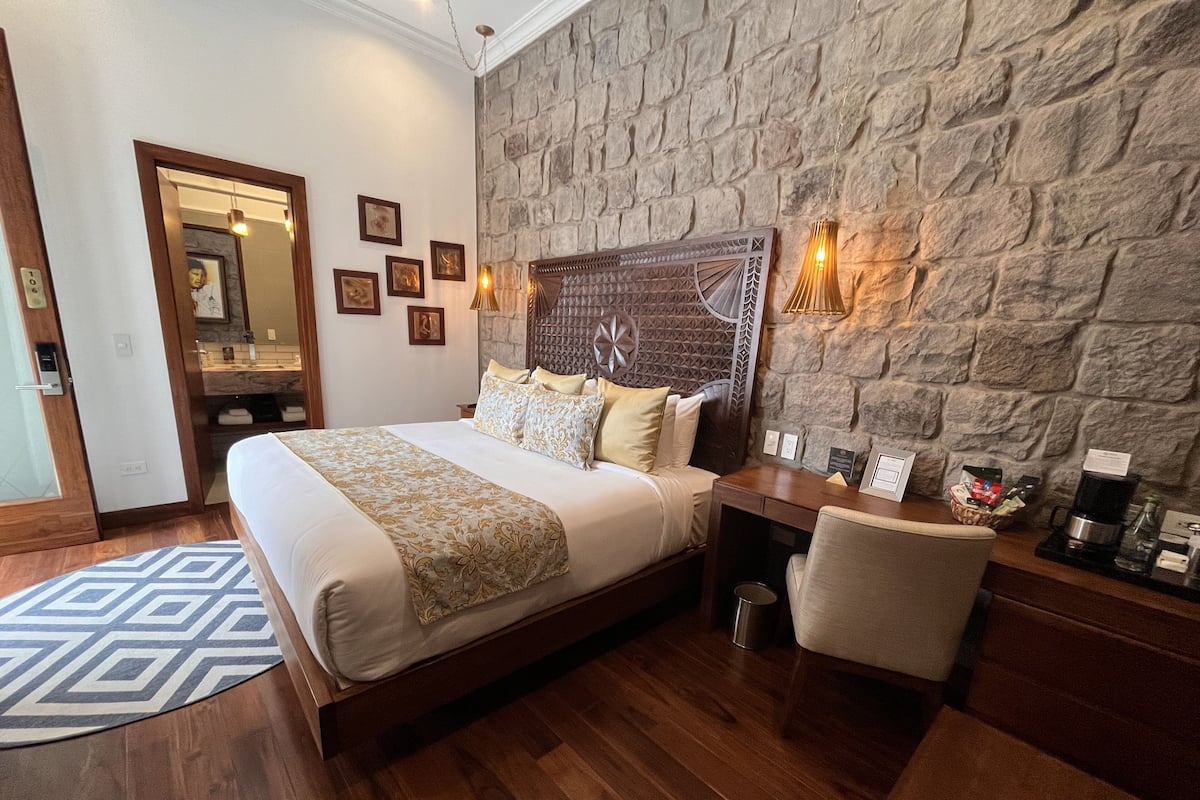 Ein Hotelzimmer mit einem großen Bett, einem hölzernen Kopfteil, einer Steinwand, einem kleinen Tisch mit Stuhl und einem durch eine offene Tür einsehbaren Badezimmer.