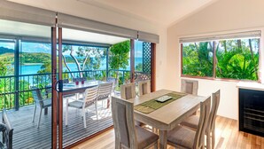 Comedor con mesa y sillas de madera, ventanas amplias con vista al mar y vegetación, y una puerta corredera de vidrio que da a una terraza al aire libre.