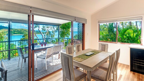 Comedor con mesa y sillas de madera, ventanas amplias con vista al mar y vegetación, y una puerta corredera de vidrio que da a una terraza al aire libre.