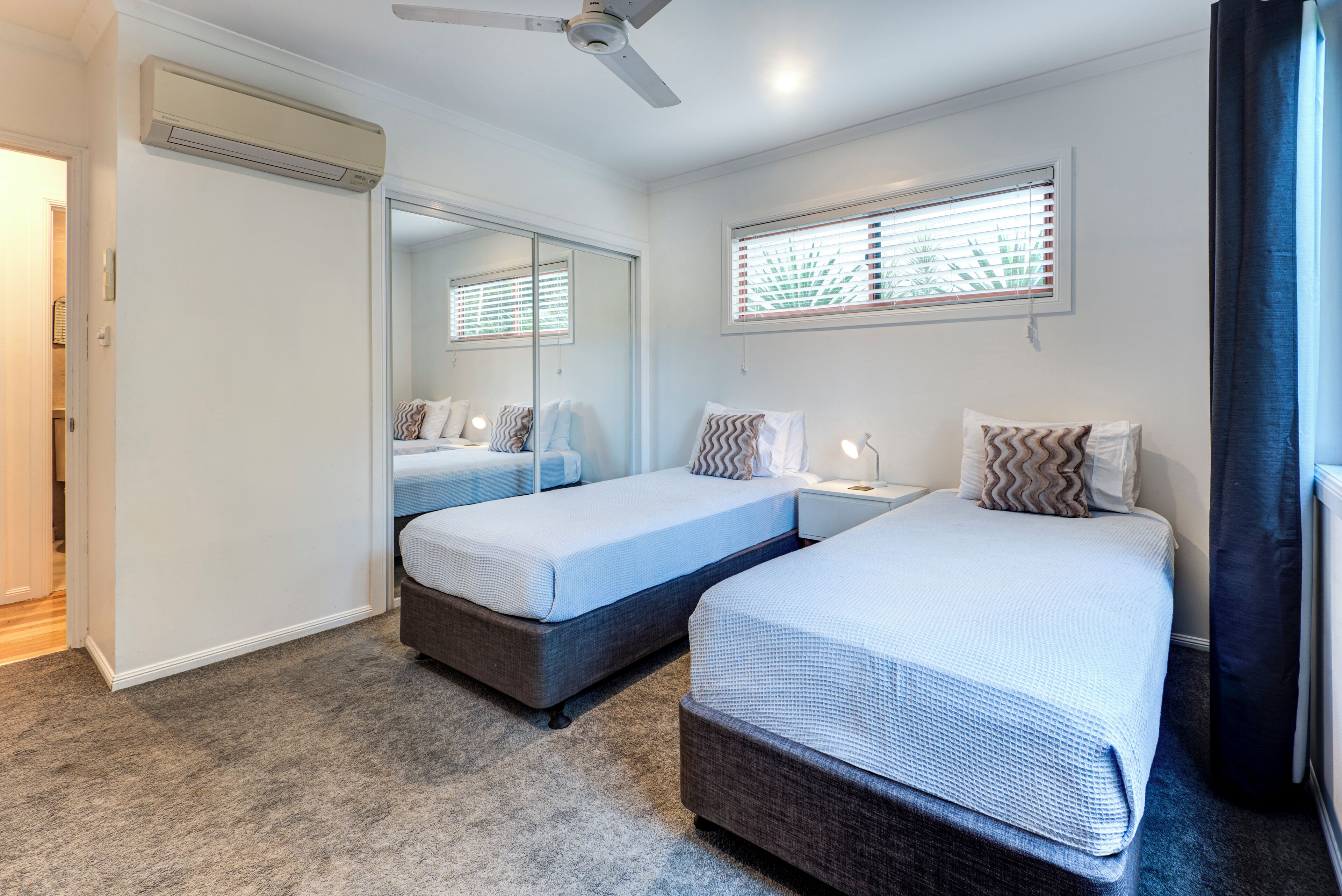 A bedroom with two single beds, a ceiling fan, an air conditioning unit, a window with blinds, and a mirror.