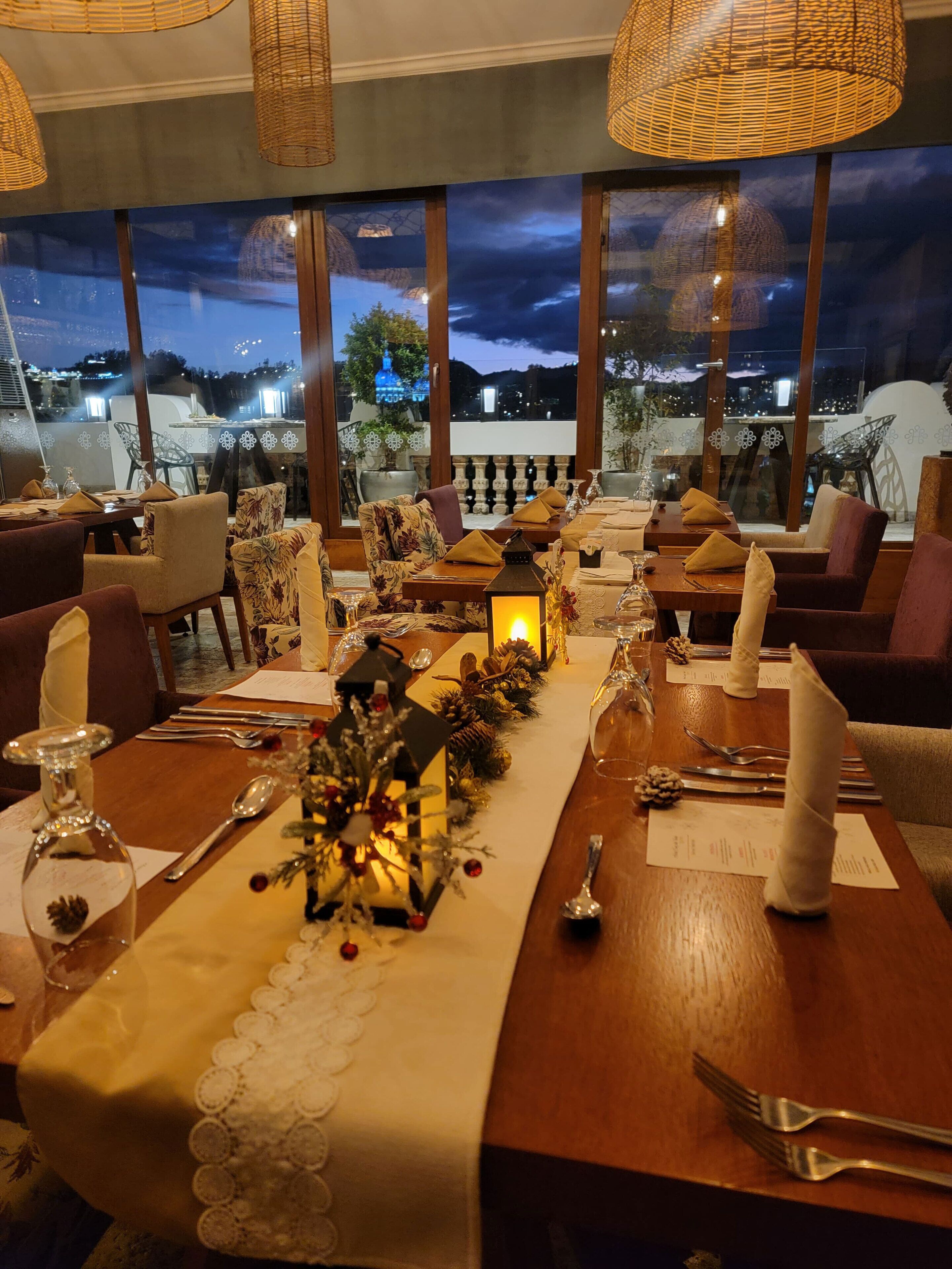 A restaurant with a long dining table set for a meal, large windows with a view of the city at night, and wicker ceiling lamps.