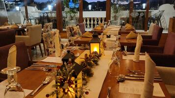 A restaurant with a long dining table set for a meal, large windows with a view of the city at night, and wicker ceiling lamps.