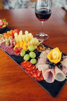 Una tabla de embutidos con diversos quesos, carnes y frutas sobre una tabla de pizarra negra, acompañada de una copa de vino tinto.