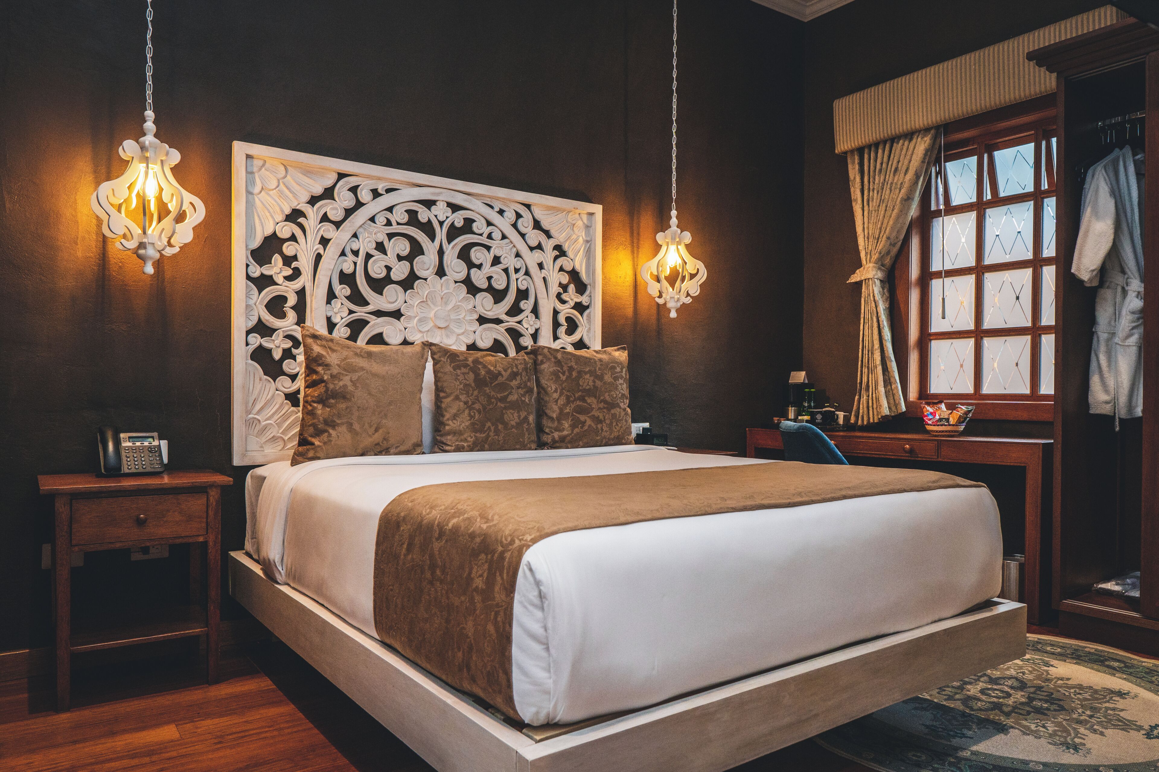 Un dormitorio con una cama grande, dos mesitas de noche, una lámpara auxiliar, un cabecero decorativo, una ventana con cortinas y una lámpara colgante.