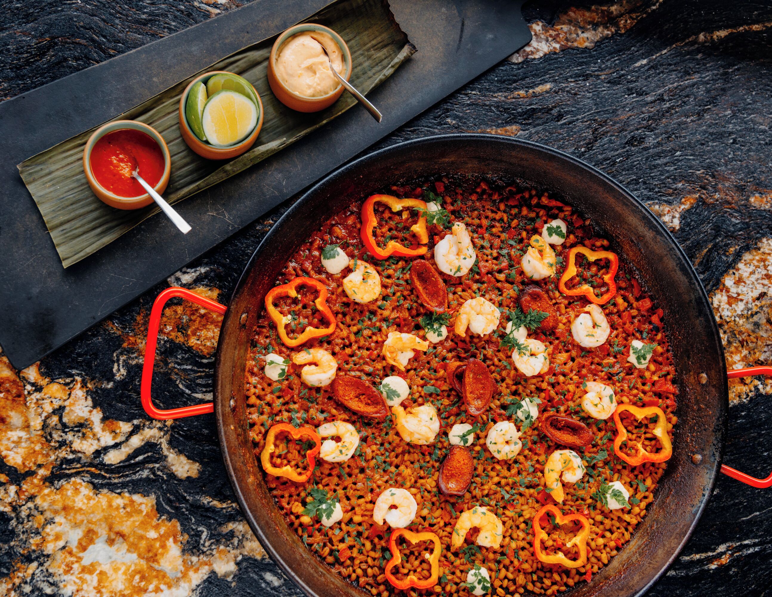 Una paella grande con gambas y pimientos, acompañada de recipientes pequeños con salsa y lima.