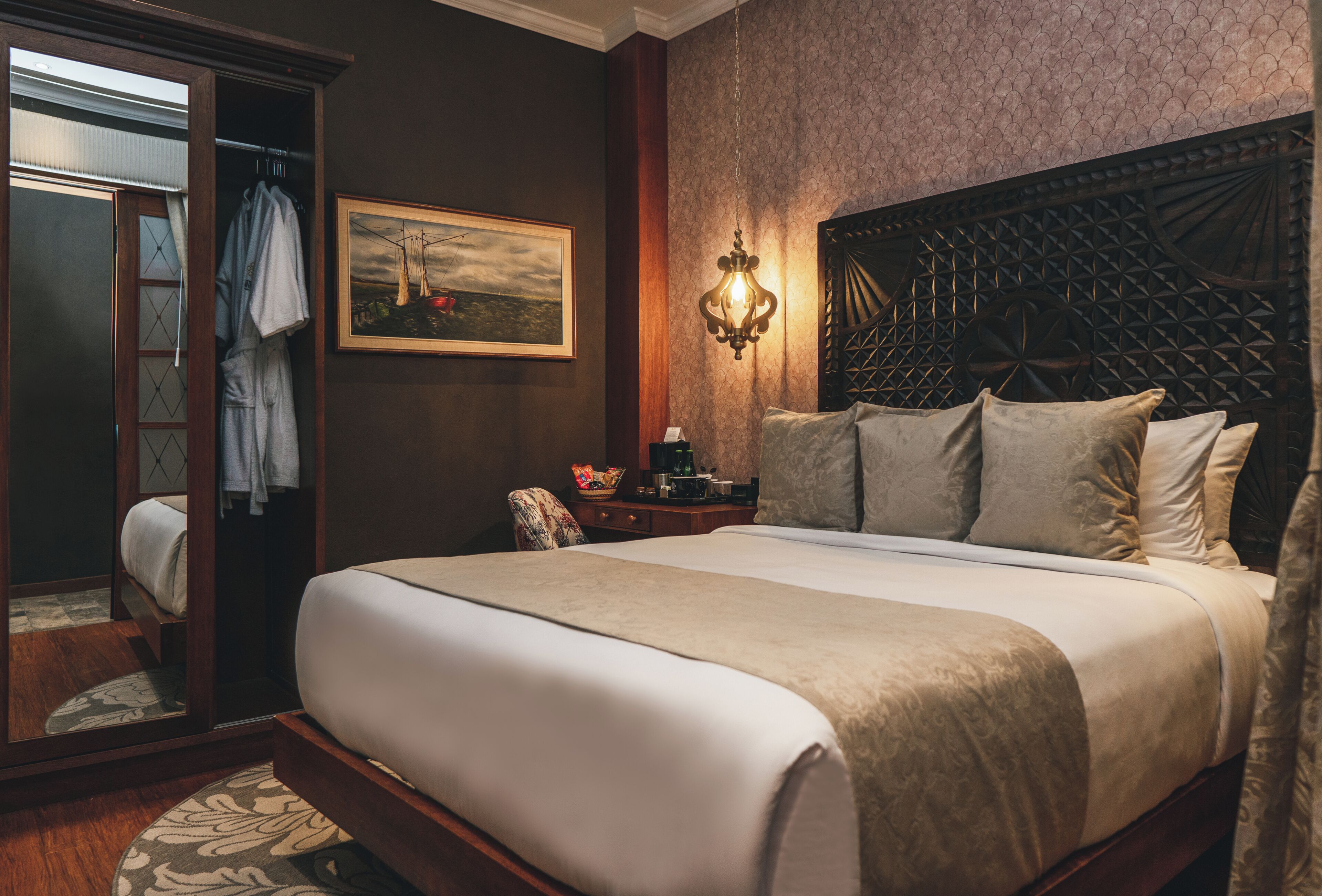A neatly made bed with a headboard, a bedside table with a lamp, a framed picture on the wall, and a door with a towel hanging on it.