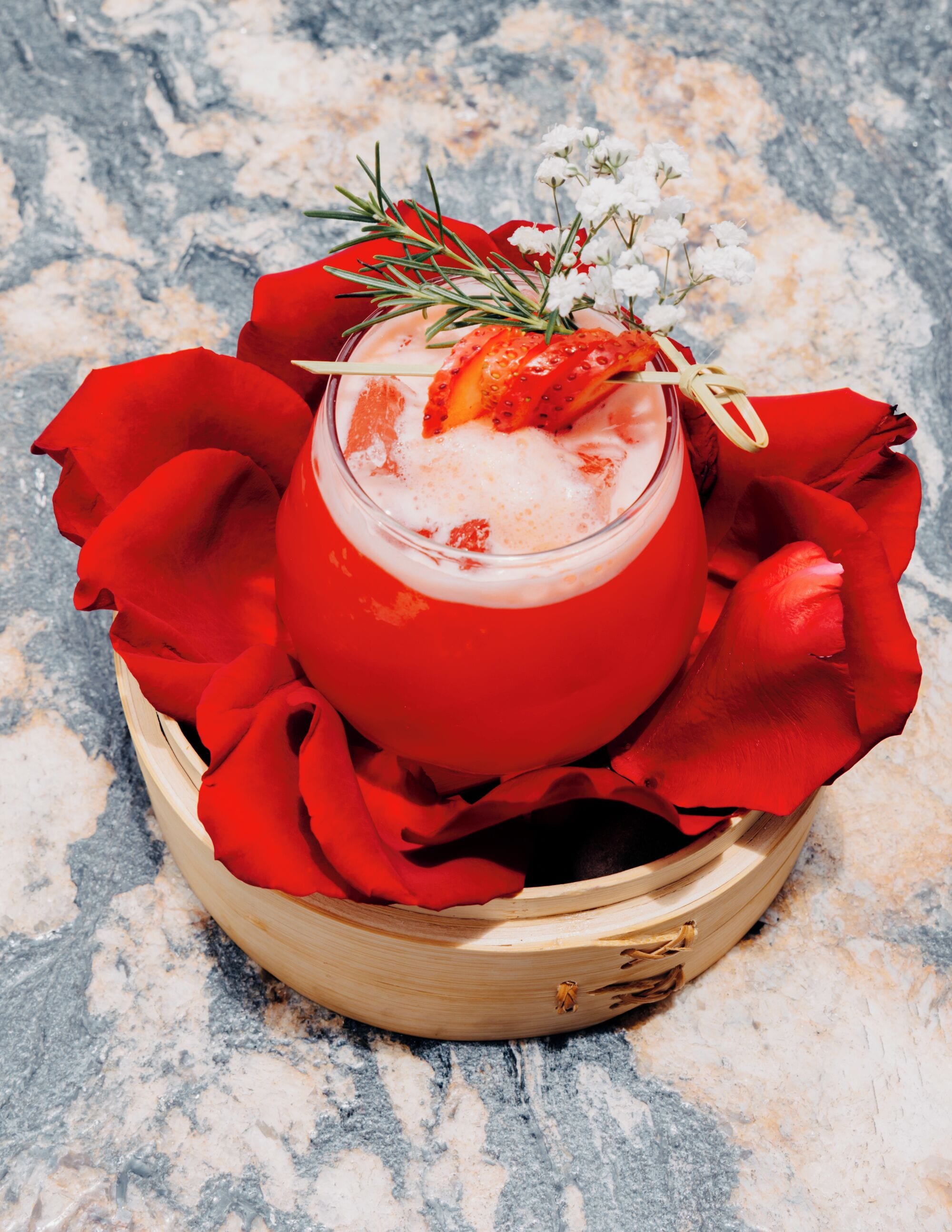 Un cóctel en un vaso rojo, adornado con fresas y romero, rodeado de pétalos de rosa roja sobre una bandeja de madera.
