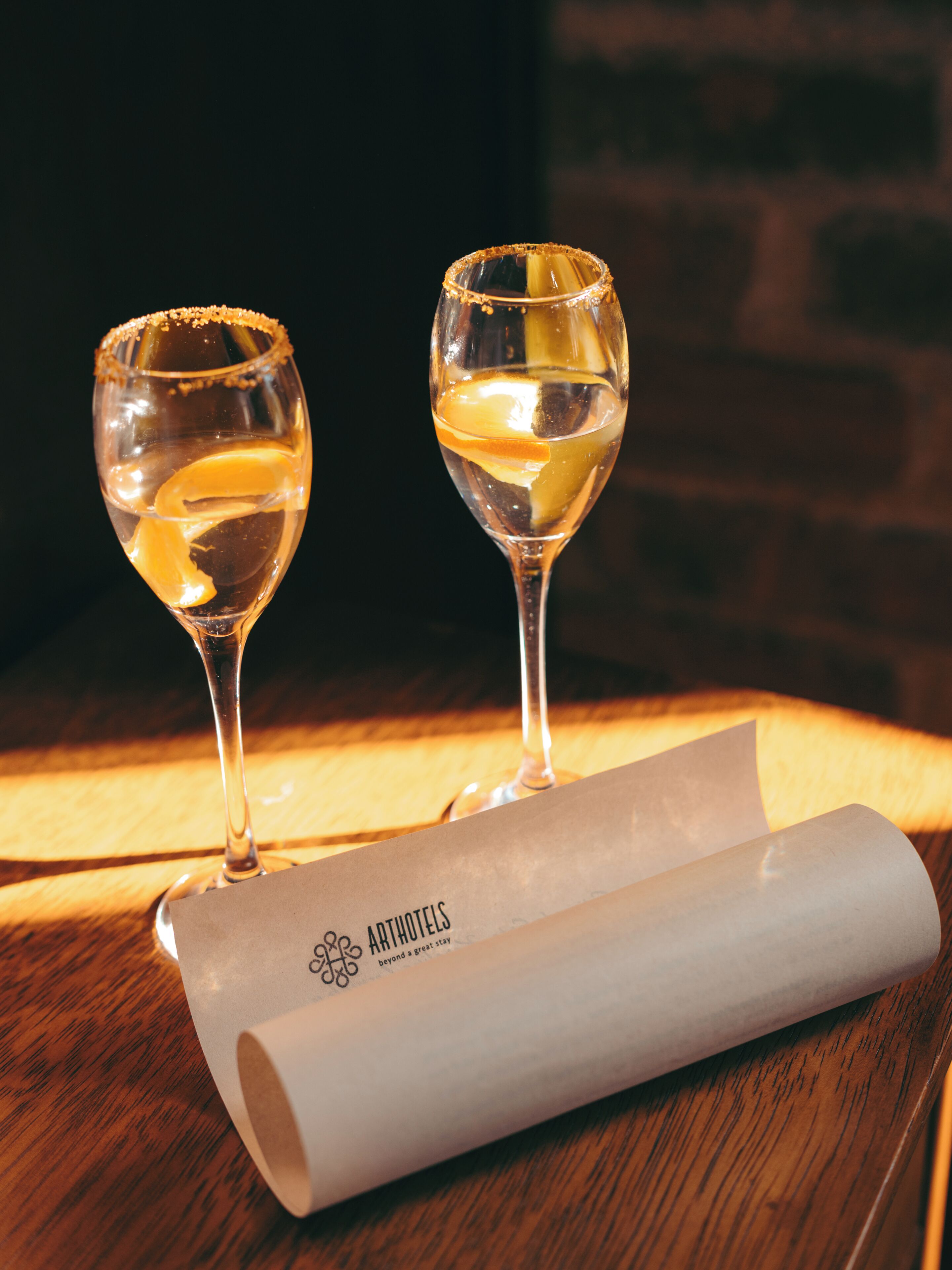 Two wine glasses on a wooden table with a rolled-up piece of paper featuring the logo 'ARTHOTELS' and the tagline 'beyond a great stay'.