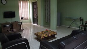 Una sala de estar con un sofá, una mesa de centro con mantel estampado, un televisor en la pared y una puerta al fondo.