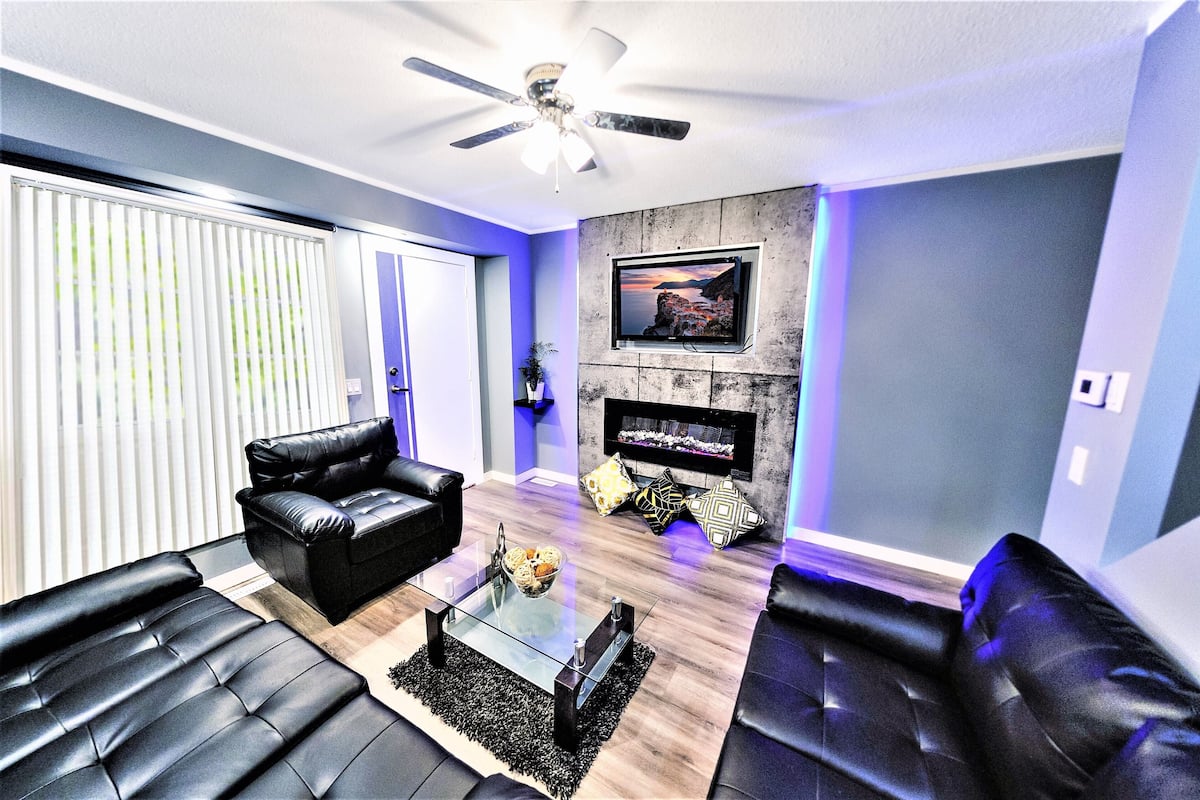 A modern living room with black leather sofas, a glass coffee table, a ceiling fan, a fireplace, and a TV mounted on the wall.