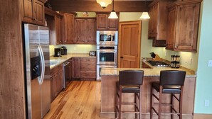 A kitchen with wooden cabinets, stainless steel appliances, granite countertops, and a bar area with stools. - Panoramic Long Range Mountain Views on 4ac, Hot Tub, Pool Table, Arcade, Firepit (West Jefferson)