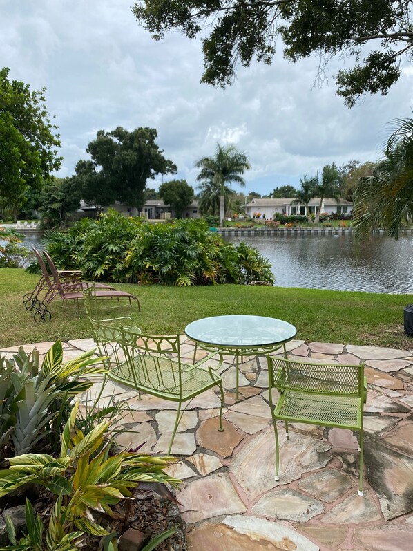 A patio with a glass table and green metal chairs, a stone pathway, and a view of a lake with houses in the background.