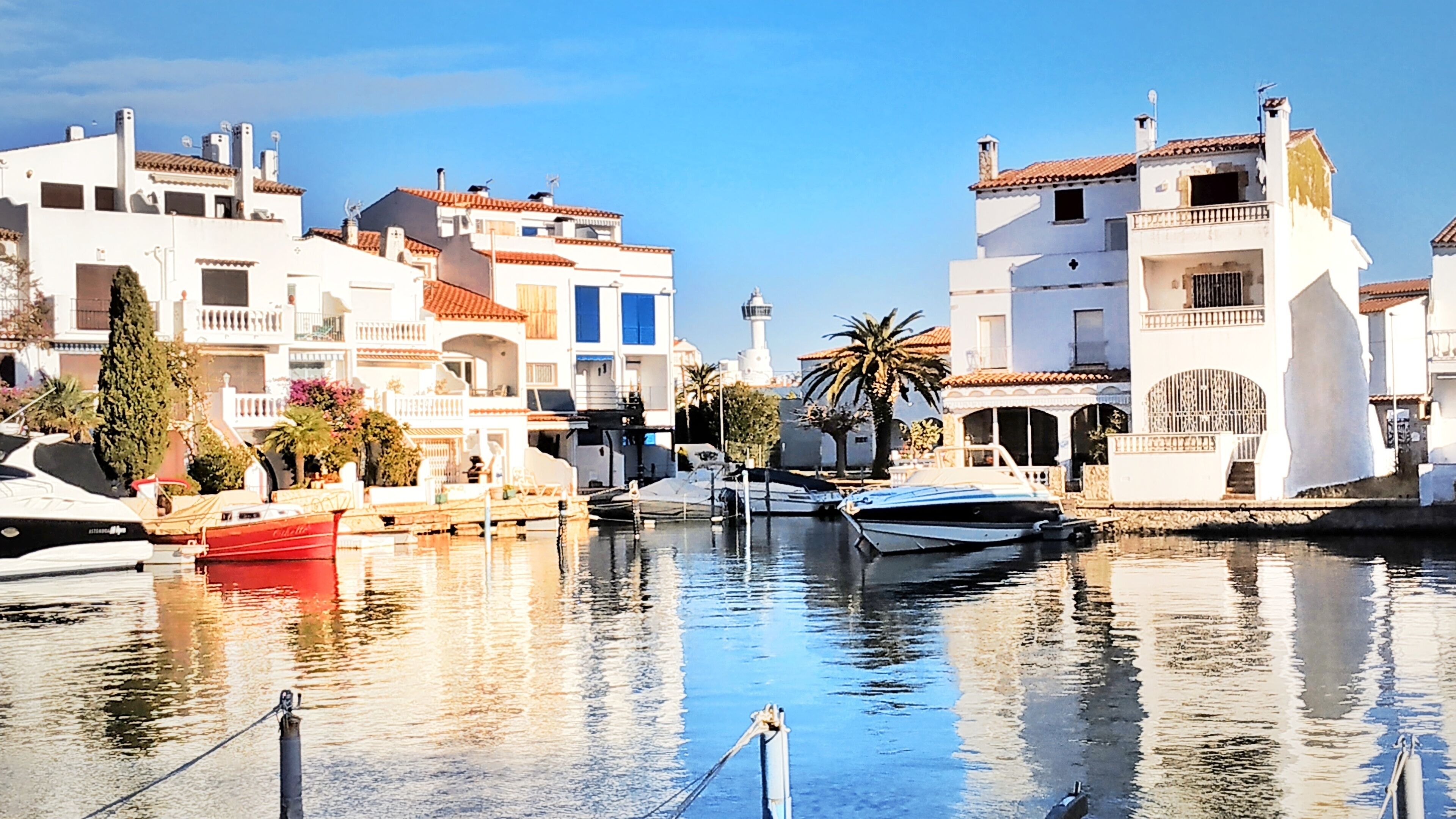A marina with boats docked, white buildings with red-tiled roofs, and a lighthouse in the background.