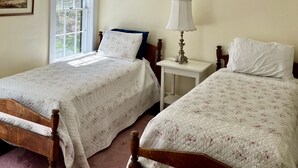 Een slaapkamer met twee eenpersoonsbedden, een nachtkastje met een lamp, een raam met zonwering en een lijst met een afbeelding aan de muur.