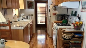 Een keuken met houten kastjes, een fornuis, een spoelbak en een eethoek met een tafel en stoelen. Er is een raam met jaloezieën, een hangklok en een klein plankje met boeken en een mand.