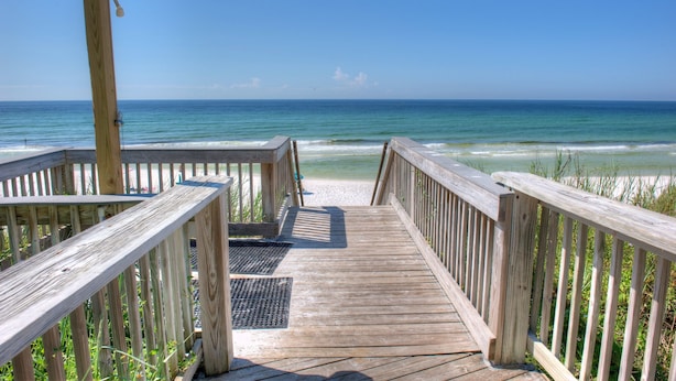 Une promenade en bois qui mène à une plage où l’eau est d’un bleu limpide et le rivage est sablonneux.