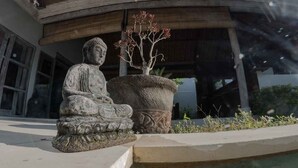 Une statue de Bouddha en pierre, placée dans un jardin, avec un grand bac à fleurs en pierre et un bâtiment en arrière-plan.