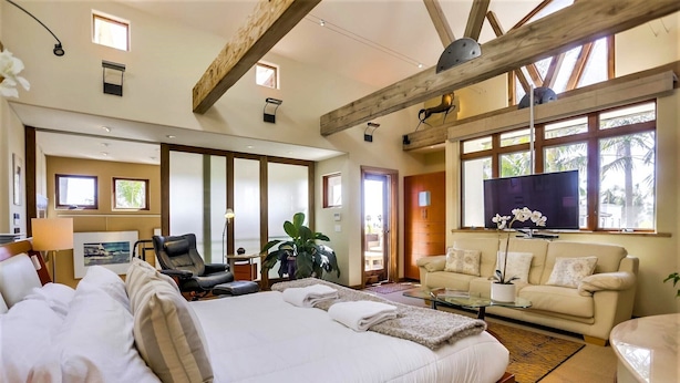 A spacious bedroom with a large bed, a sofa, a TV, and a window with a view of palm trees. - Tranquility La Jolla | Custom Home w/ Rooftop (San Diego)