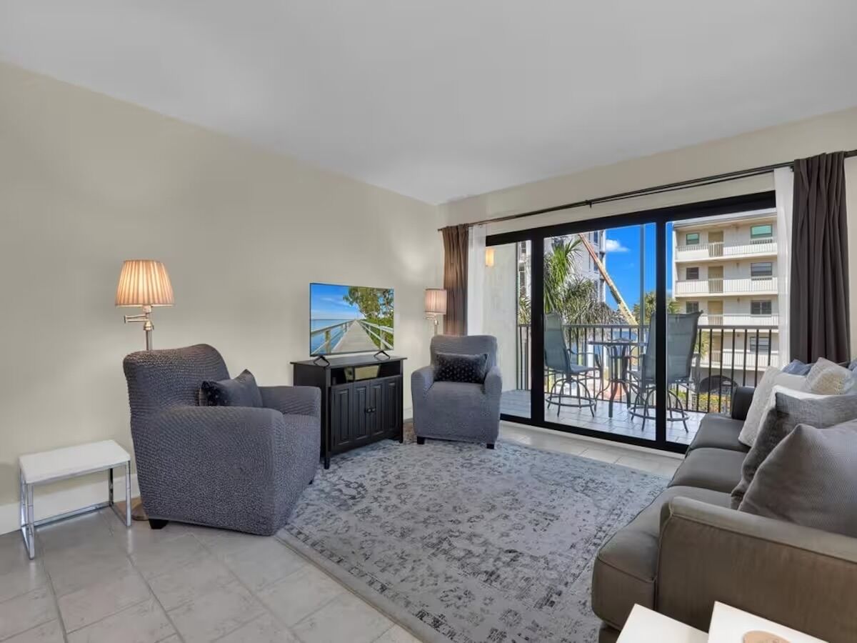 A modern living room with a grey sofa set, a small white side table, a dark wooden cabinet, and a large window offering a view of a balcony and buildings.