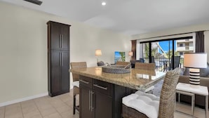A modern kitchen with dark cabinetry, a granite countertop, and a dining area with wicker chairs and a small table. - Walk to Beach & Rooftop Pool by Luxury Resorts (Naples)