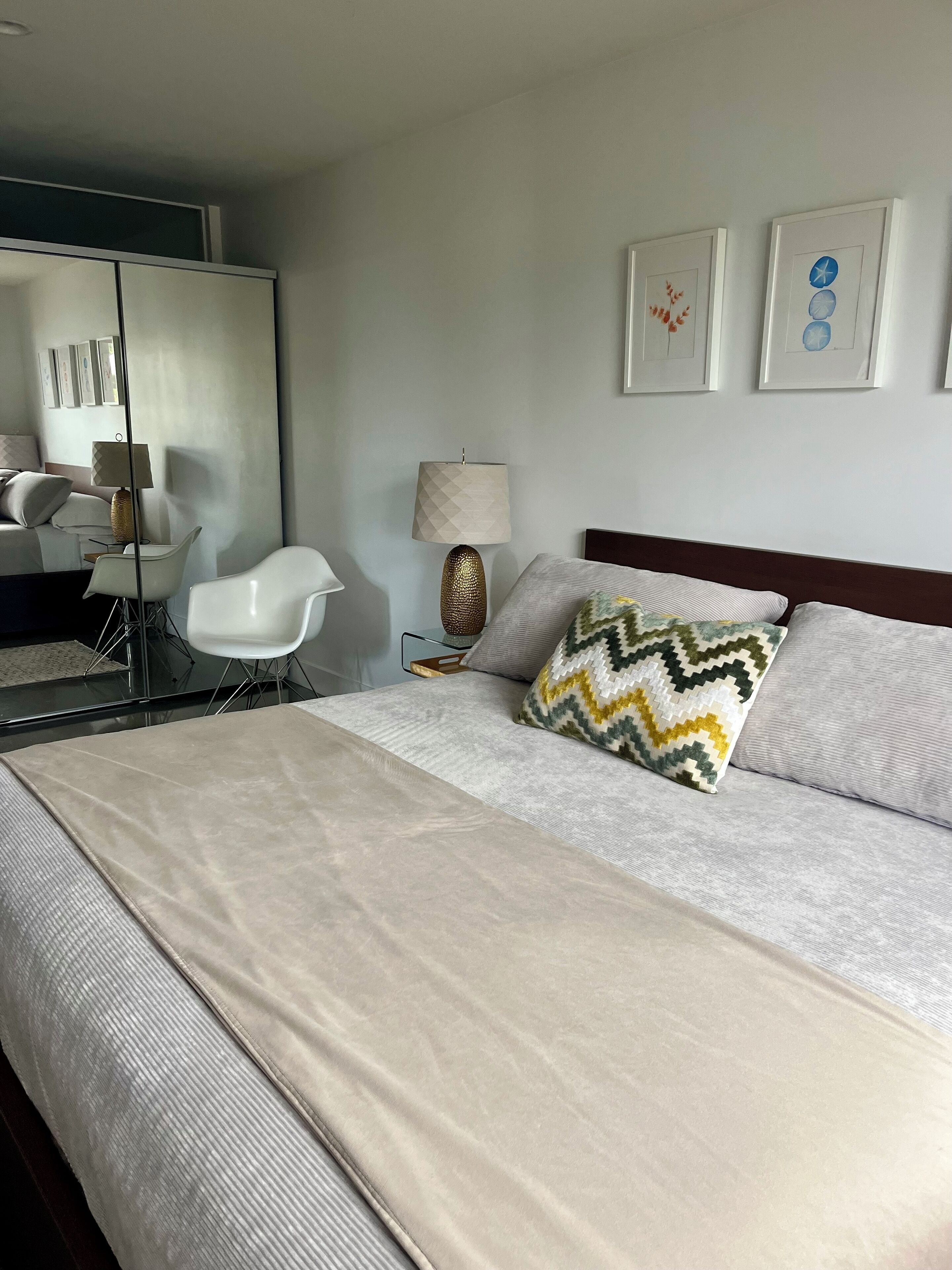 Quarto de hotel moderno com uma cama grande, uma mesinha de cabeceira, uma luminária, uma cadeira e um espelho.
