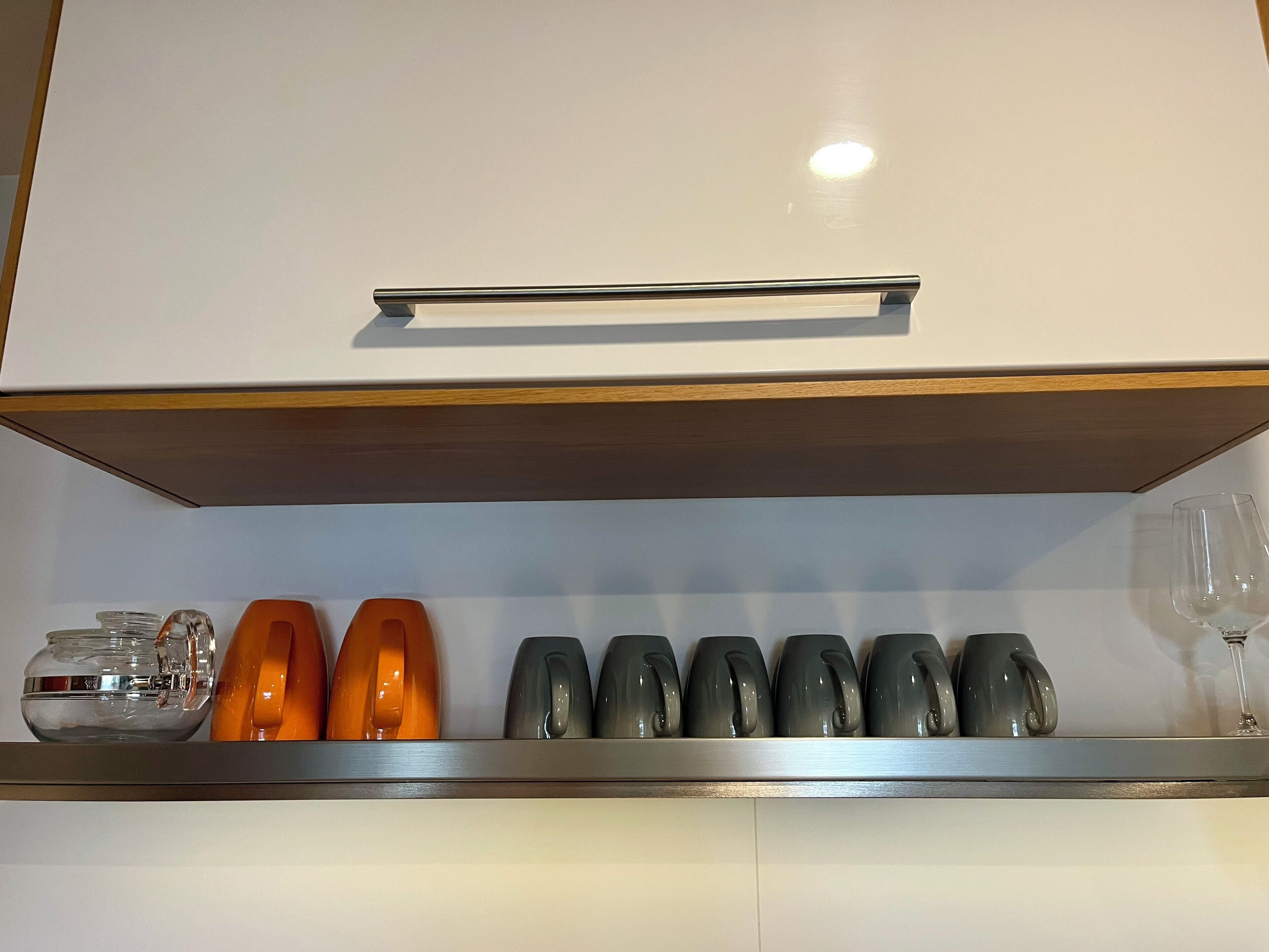 Una cocina con un armario blanco, una repisa con tazas variadas, un tarro de vidrio y una copa de vino.