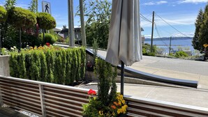 Eine Uferpromenade mit einer gebogenen Bank, einem Blumenbeet mit gelben Blumen und Blick auf das Wasser sowie hügelige Landschaft in der Ferne.