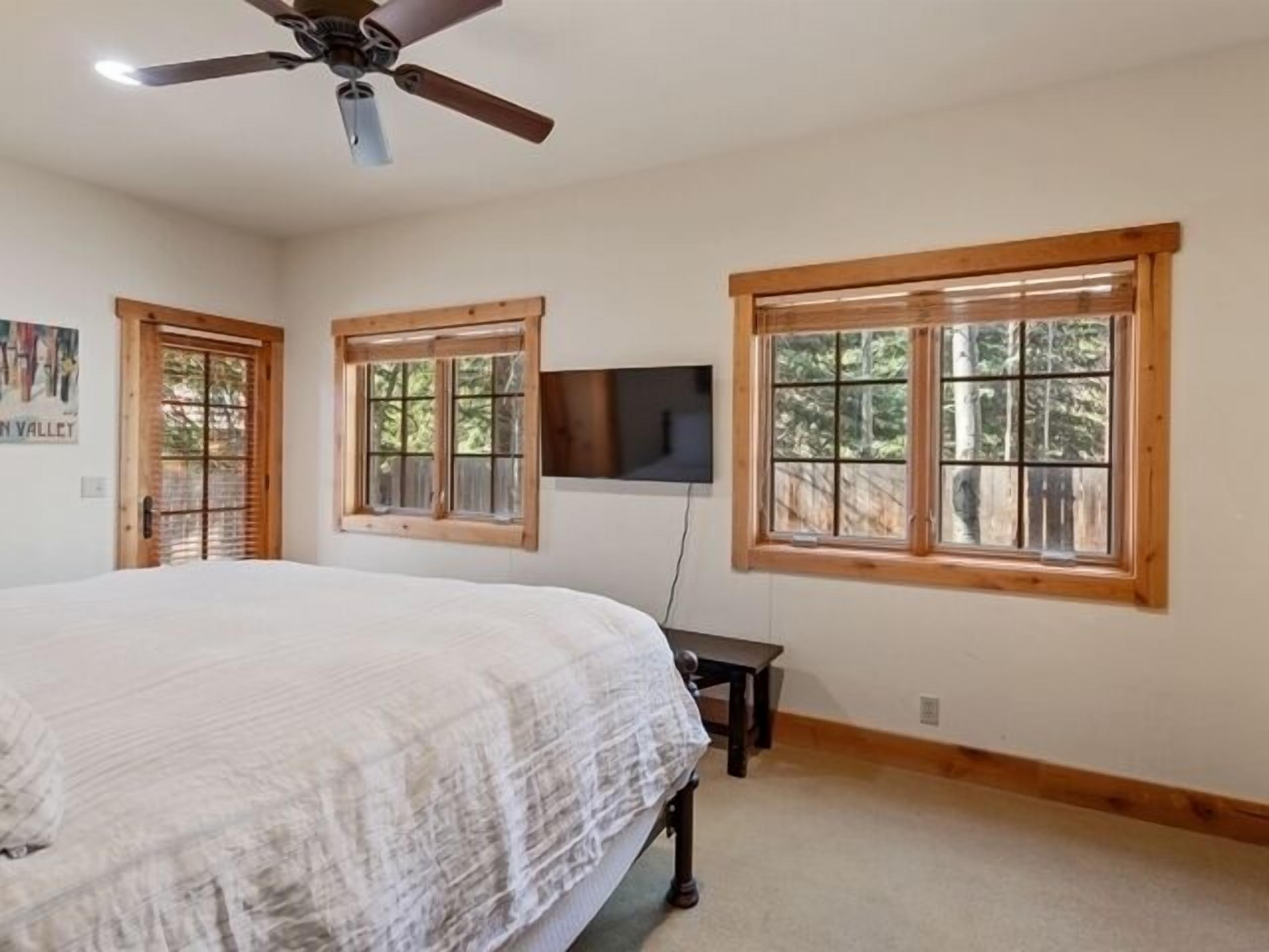 A bedroom with a bed, a ceiling fan, a TV mounted on the wall, and two windows with wooden frames.