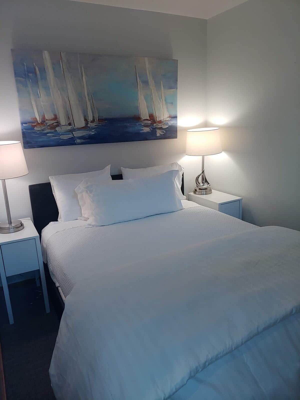 A neatly made bed with white linens, two bedside lamps, a painting of sailboats on the wall, and a small nightstand with a lamp.