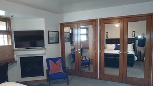 A hotel room with a bed, a TV, a fireplace, a blue chair, a wardrobe, and a mirror.