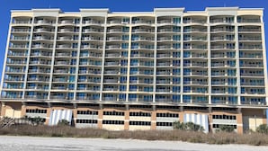 Un hôtel à plusieurs étages, doté de nombreux balcons, d’un toit plat et bordé d’une plage de sable au premier plan.