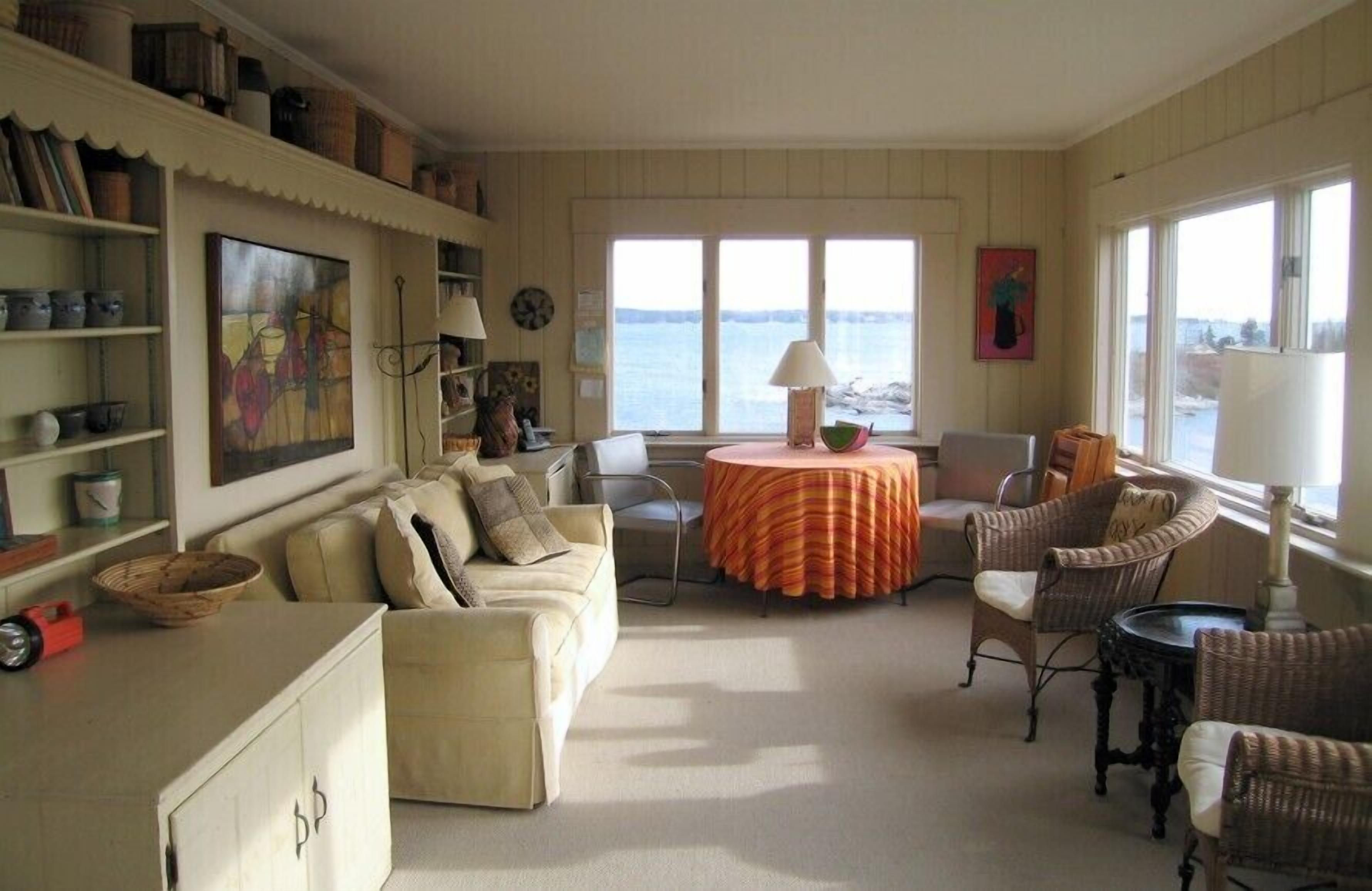 Un salon chaleureux avec un canapé beige, des fauteuils en osier, une table ronde recouverte d’une nappe à rayures et une vue sur la mer par de grandes fenêtres.
