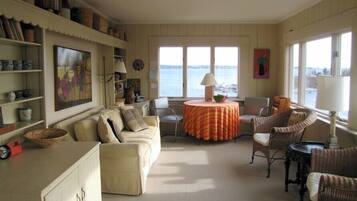 Un salon chaleureux avec un canapé beige, des fauteuils en osier, une table ronde recouverte d’une nappe à rayures et une vue sur la mer par de grandes fenêtres.