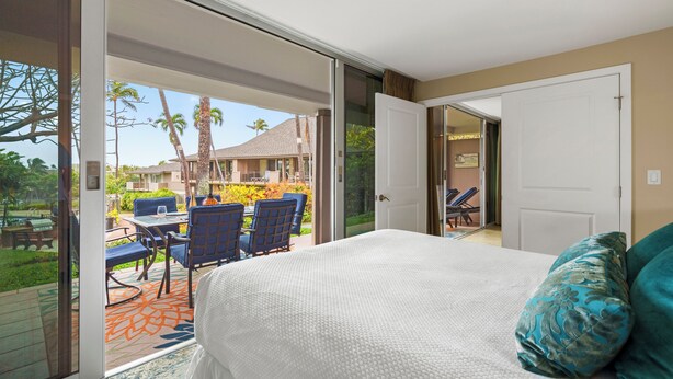 Ein Schlafzimmer mit einem großen Bett, einer Schiebetür, die auf eine Terrasse im Freien mit Tisch und Stühlen führt, und Blick auf ein Gebäude mit einem geneigten Dach.