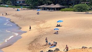 Ein Strand mit Menschen, die sich sonnen und am Ufer entlanggehen. Im Hintergrund sind Sonnenschirme und ein Gebäude zu sehen.