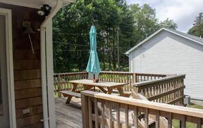 A wooden deck with picnic tables and a covered umbrella, a small shed, and a house in the background. - Charming & spacious beach home in Bonnet Shores, Narragansett (Narragansett)