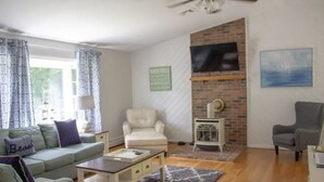 A living room with a brick fireplace, a ceiling fan, a television mounted on the wall, and a large window with patterned curtains. - Charming & spacious beach home in Bonnet Shores, Narragansett (Narragansett)