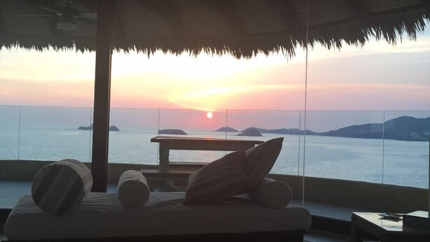 A sunlit room with a bed, pillows, and a table, overlooking a body of water and distant islands at sunset. - Los mejores atardeceres en Ixtapa Zihuatanejo (Ixtapa)